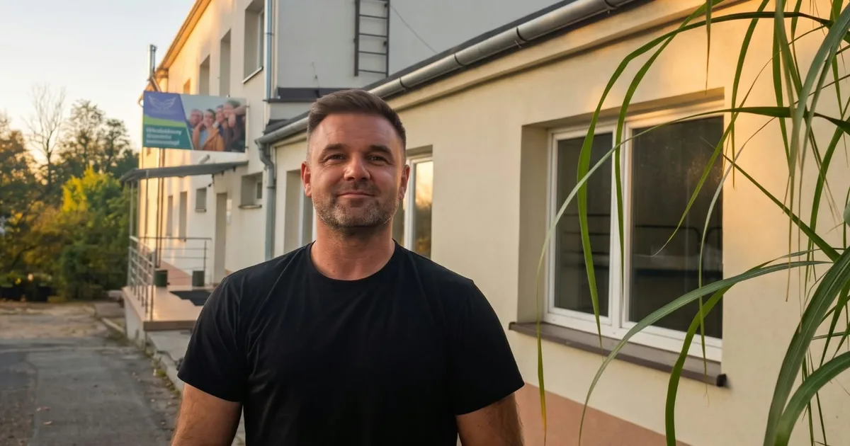 Dawid Mikosiński w czarnej koszulce stoi przed jasnym budynkiem, w tle zieleń i światło zachodzącego słońca.